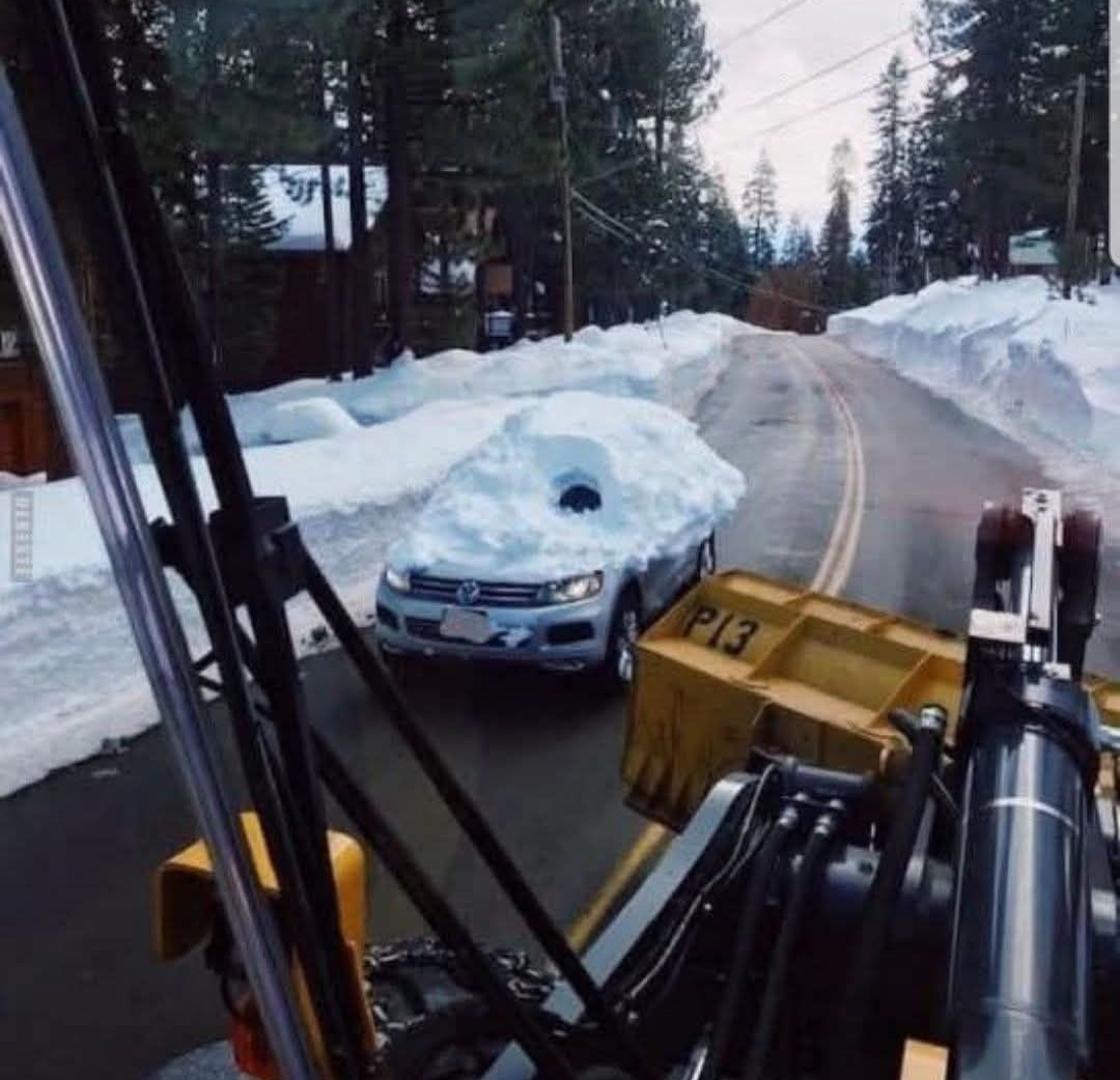 Auto pokryté snehom na horskej ceste vedľa snežného pluhu, obklopené zasneženou krajinou.
