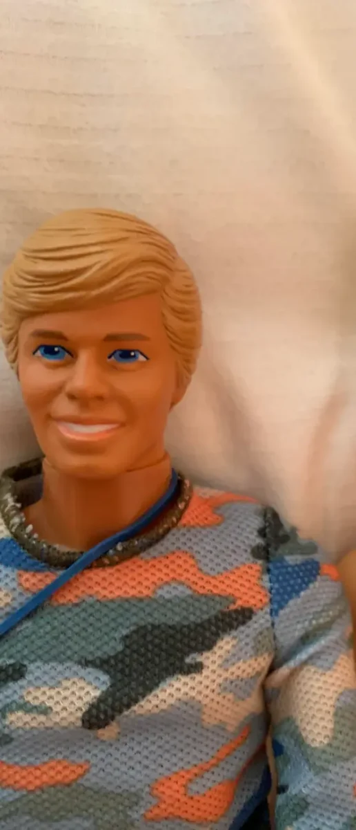 Bábika v maskáčovom oblečení s blond vlasmi a modrými očami, zblízka.