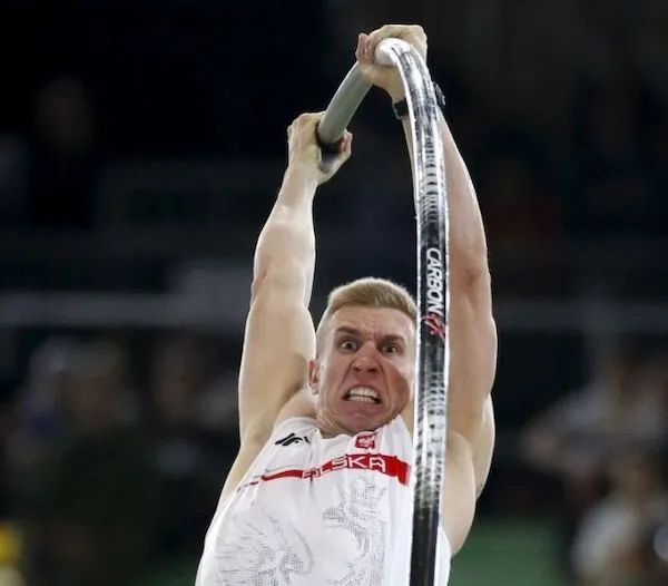 Športovec v bielom drese počas skoku o žrdi na atletickej súťaži.