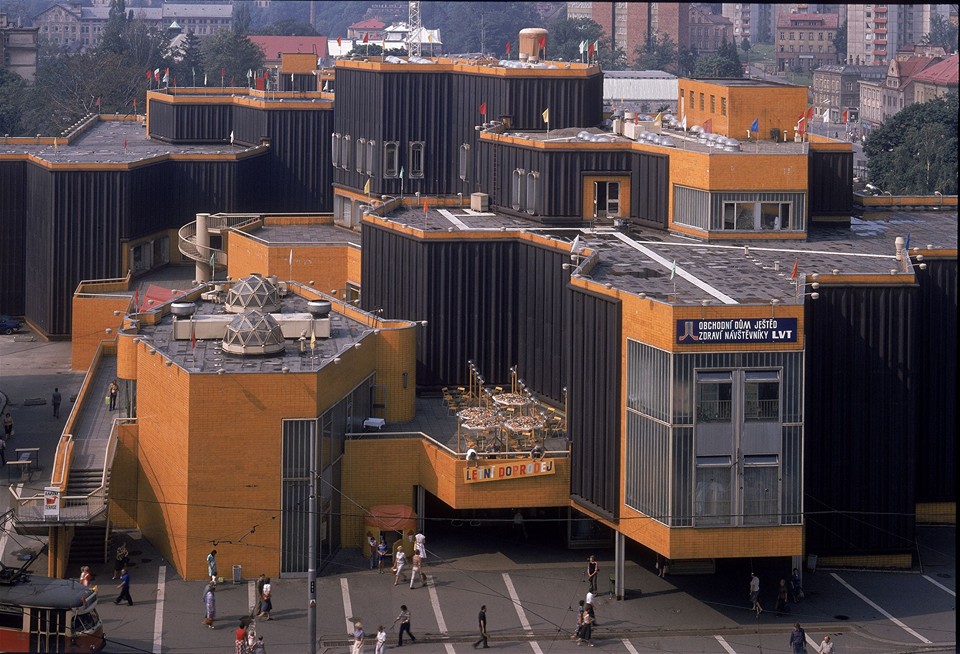 Moderná budova s unikátnou architektúrou a ľuďmi na ulici. Tehly a čierne panely dominujú exteriéru stavby.