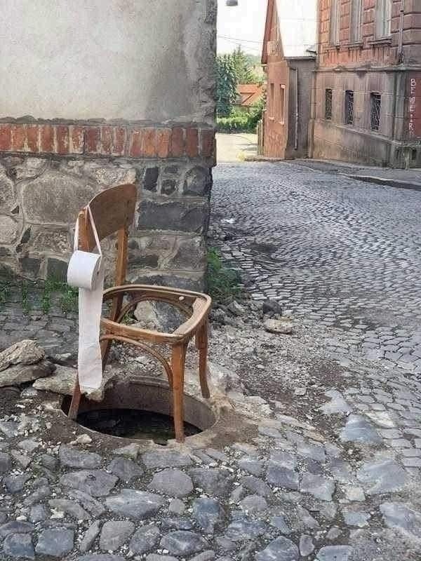 Stolička nad otvorom na dláždenej ulici so zaveseným toaletným papierom, vzbudzuje dojem improvizovanej verejnej toalety.
