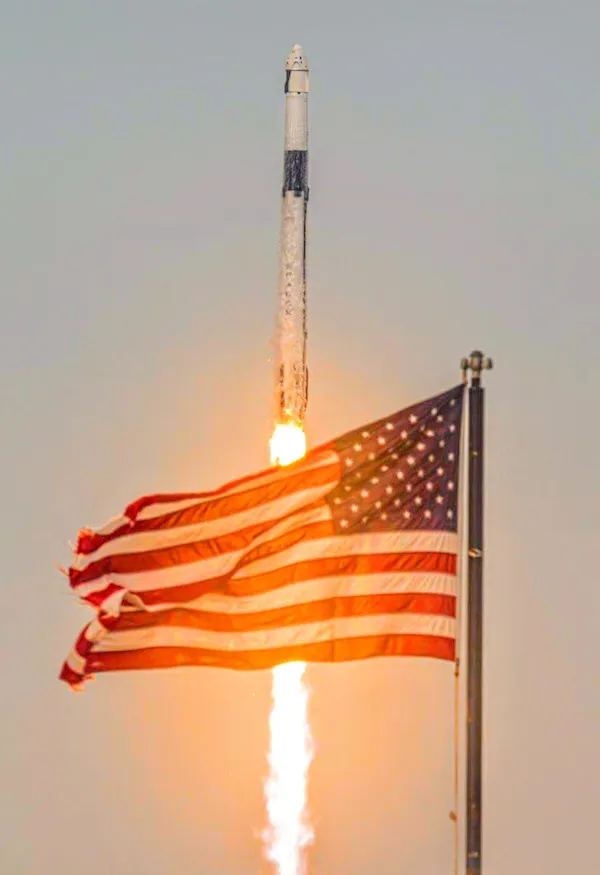 Raketa odlieta do vesmíru za americkou vlajkou, vznášajúc sa nad obzorom pri západe slnka.