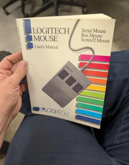 Používateľ drží starý manuál k Logitech sériovej myši s farebnými prúžkami na obale.