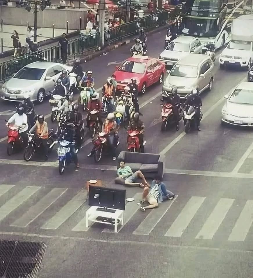 Skupina ľudí relaxuje na gauči uprostred prechodu, zatiaľ čo okolo premávajú autá a motocykle. Nepredvídateľná situácia na ceste.