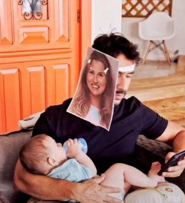 Otec dojčí dieťa s vtipnou fotografiou ženy na tvári, sedí na gauči a používa telefón.