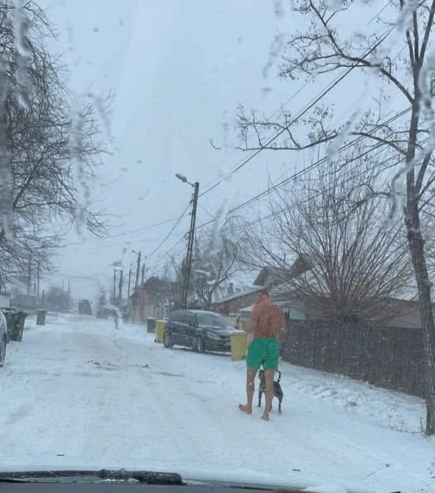 Muž bez trička so psom na zasneženej ulici počas zimného dňa.