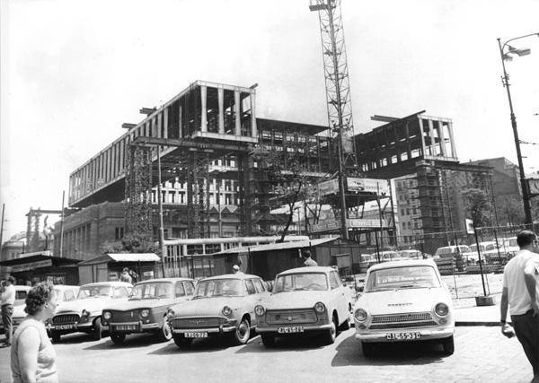Historická fotografia stavby s autami v popredí, žeriavom a rozostavanou budovou v centre, 20. storočie.