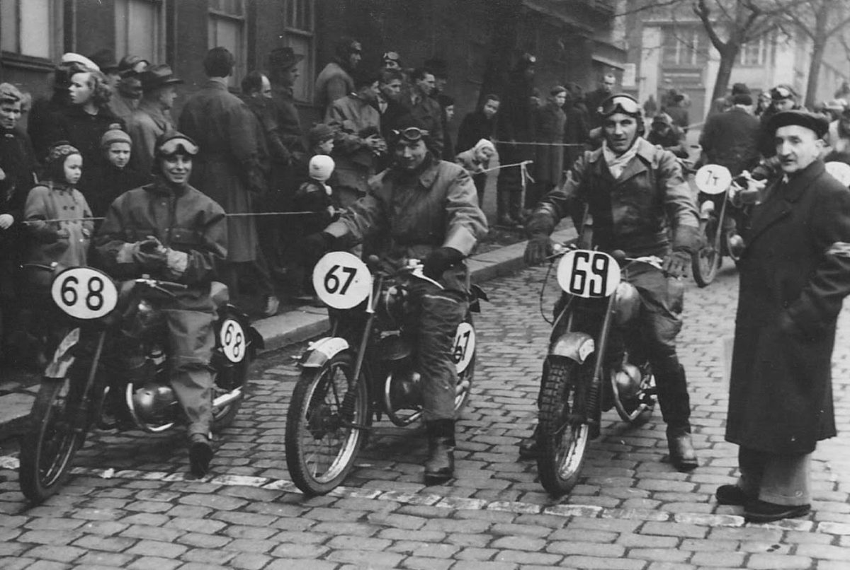Pretekári na historických motocykloch pred štartom, obklopení davmi divákov na dláždenej ulici.