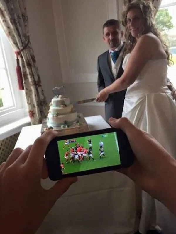 Nevesta a ženích krájajú svadobnú tortu, zatiaľ čo pozornosť je na mobile s rugby zápasom.