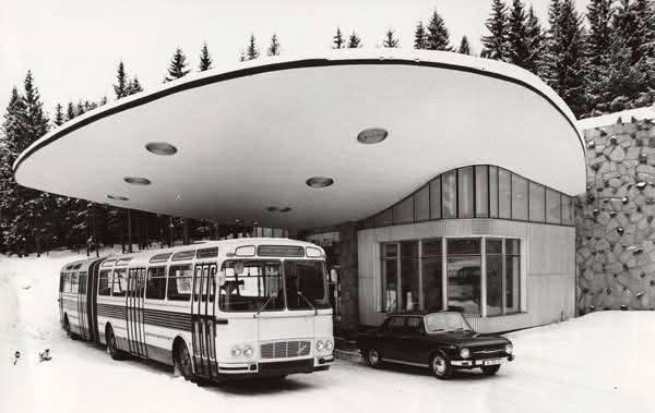 Retro autobus a auto pred modernistickou stanicou v zimnej krajine so zasneženými stromami na pozadí.