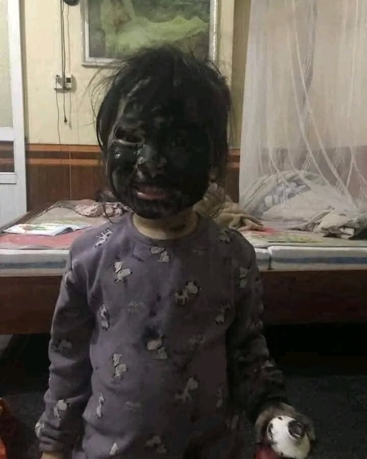 Dieťa so zašpinenou tvárou v pyžame stojí pred posteľou, držiac malú hračku v ruke.
