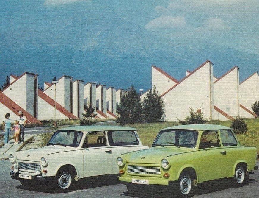Dva vozidlá Trabant parkujú pred výškovými budovami s výraznou architektúrou na pozadí za slnečného dňa.