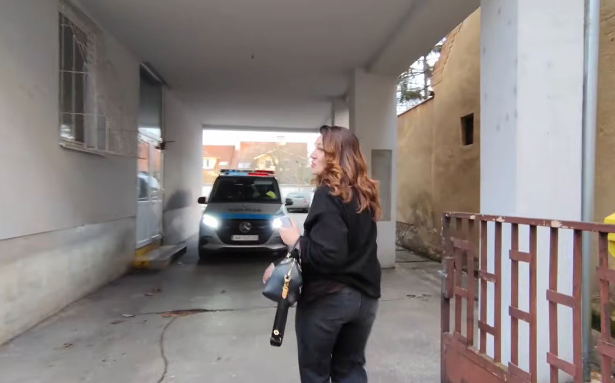 Žena stojí pri vjazde, oproti je policajné auto. Atmosféra je napnutá, lokácia mestská.