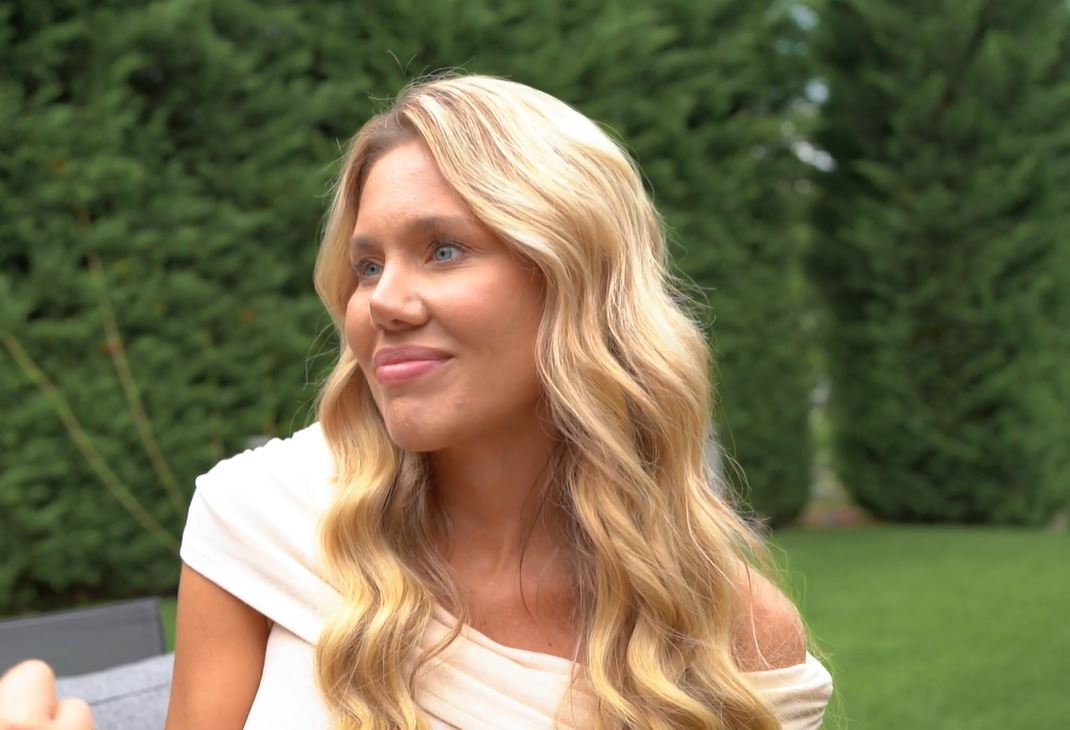 Žena s dlhými blond vlasmi sedí vonku na záhrade, oblečená v bielom, s prírodným prostredím v pozadí.