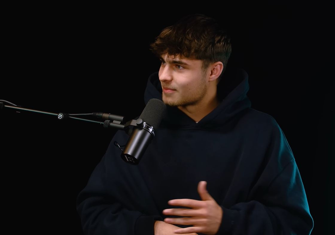 Osoba hovorí do mikrofónu počas podcastu, oblečená v tmavej mikine.