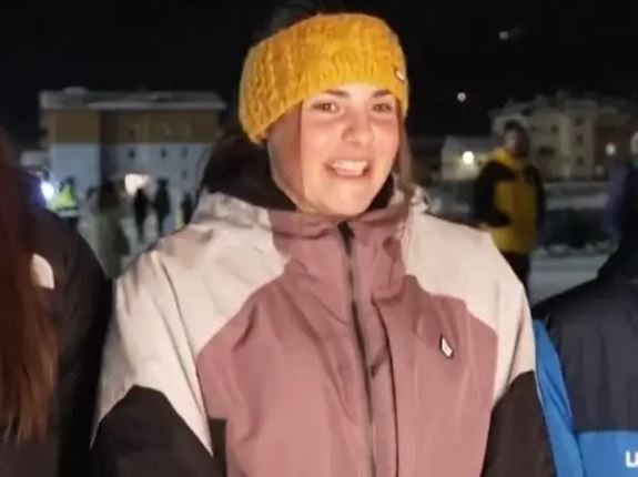 Mladá žena v teplej bunde a žltej čiapke sa usmieva počas zimného večera na pozadí osvetlenej dediny.