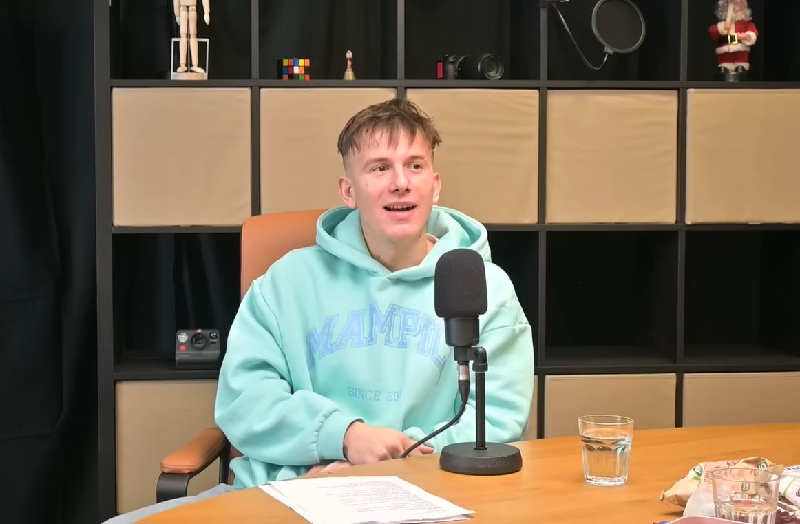 Muž v modrej mikine hovorí do mikrofónu počas podcastovej epizódy, pozadie s regálmi.