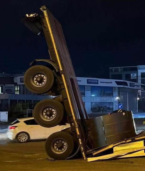 Nákladné auto vo vertikálnej polohe na ceste v noci, biely sedan prechádza okolo.