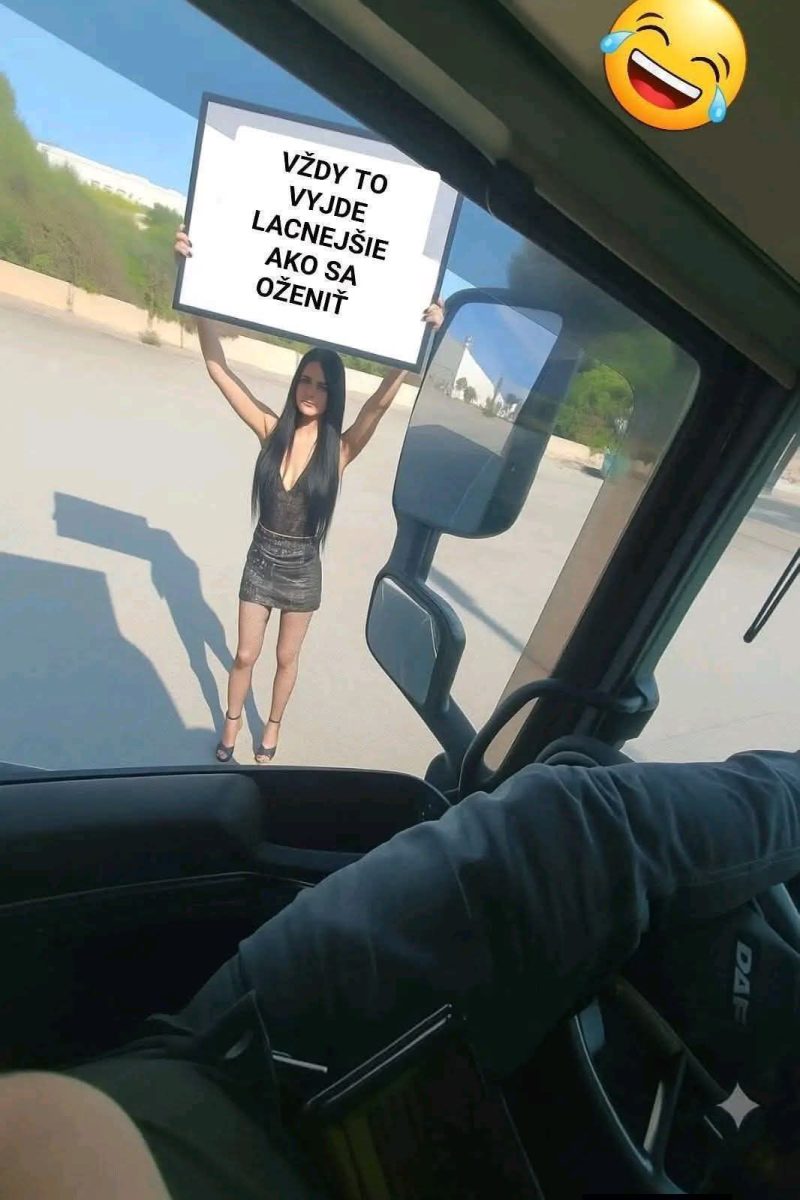 Žena drží tabuľu s vtipným nápisom pred nákladným autom, vodič sleduje scénu.