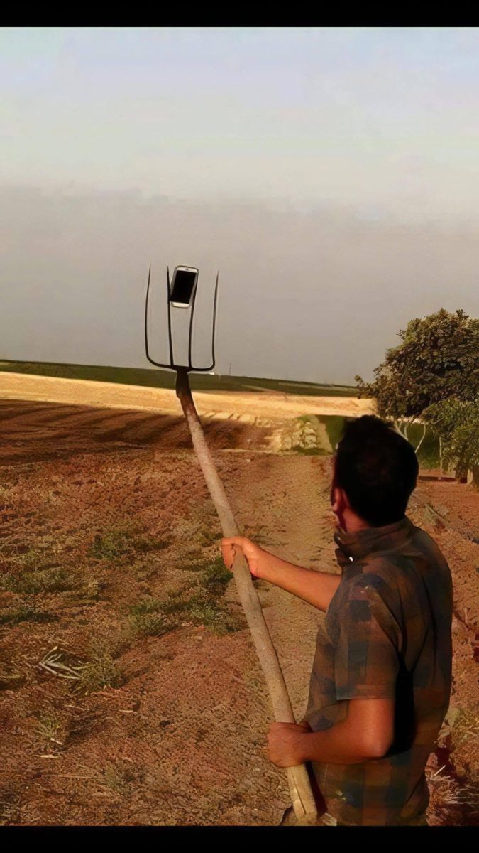 Muž drží vidly so smartfónom namiesto selfie tyče na vidieckej ceste pod modrou oblohou.