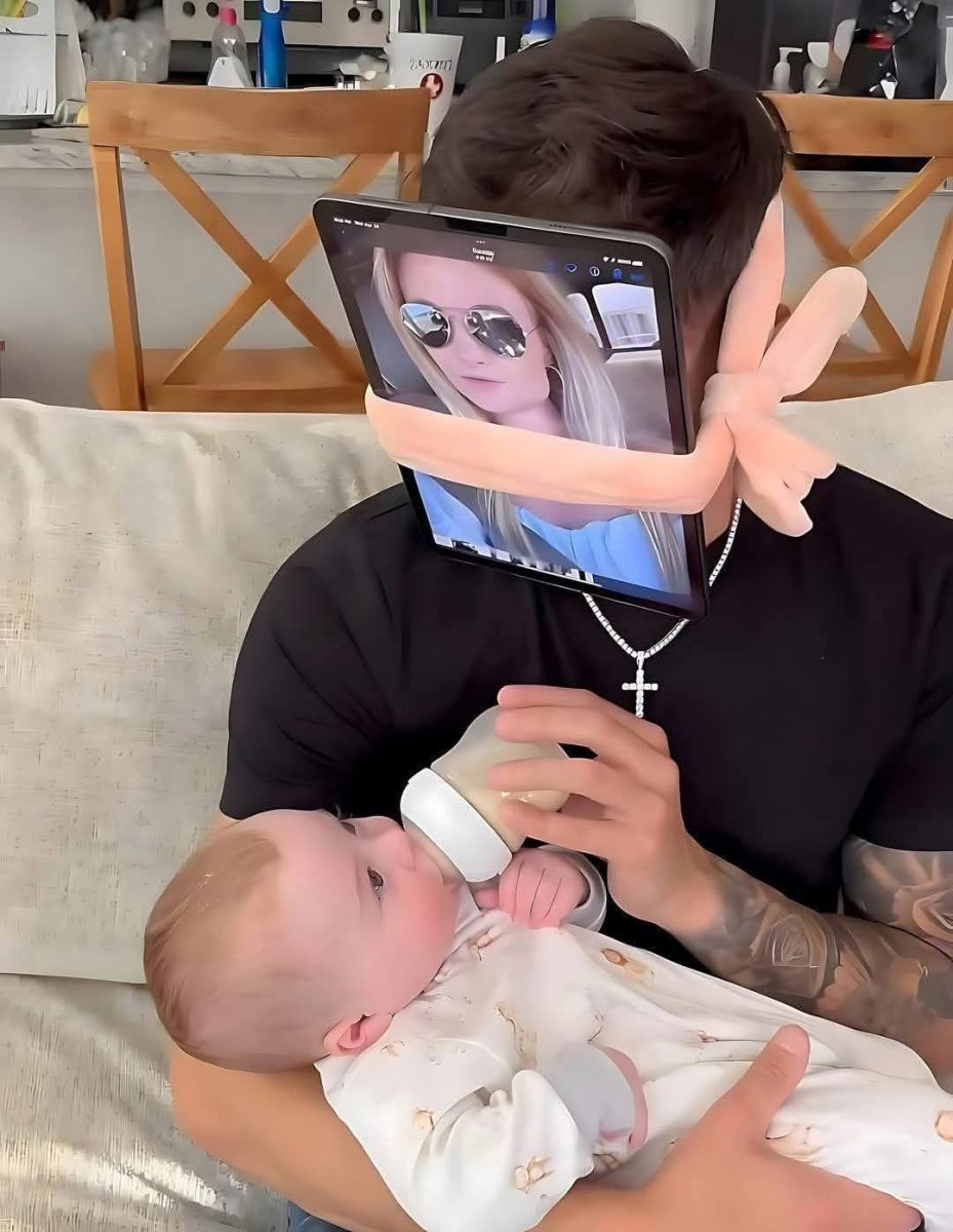 Muž dojčí dieťa, zatiaľ čo na tvári má upevnený tablet s fotografiou ženy.