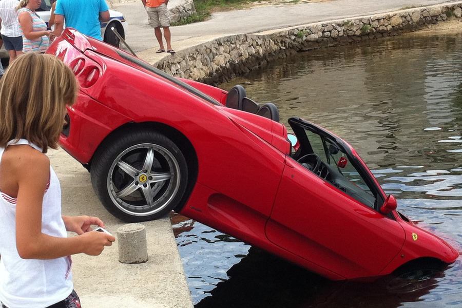 Červené športové auto napoly ponorené vo vode pri brehu, okolo stojí skupina ľudí.