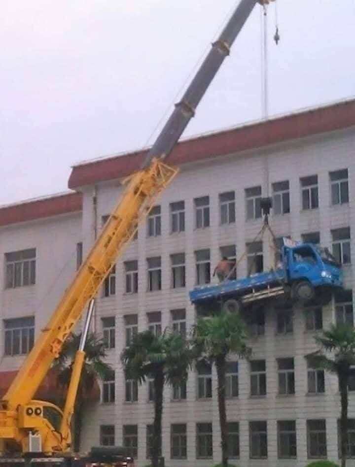 Žeriav zdvíha modré nákladné auto pred budovou, čo vytvára nevšedný pohľad.