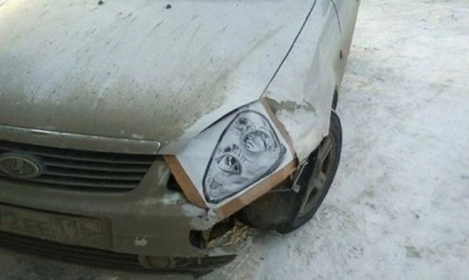 Auto s poškodeným predným svetlom opravovaným pomocou lepenky, stojí na zasneženej vozovke.