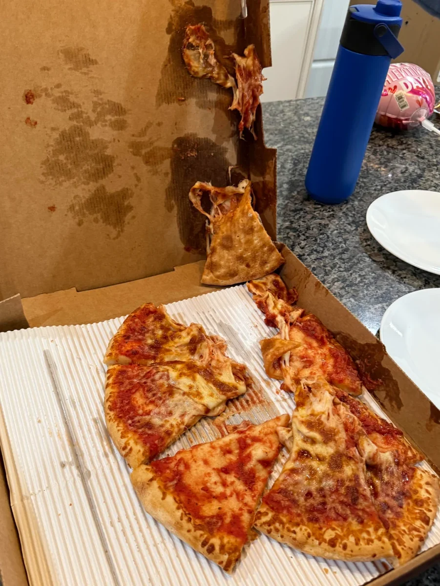 Pizza v škatuli nalepenej na vrchnák, roztečená so syrom a omáčkou na kartóne, vedľa modrej fľaše na vode.