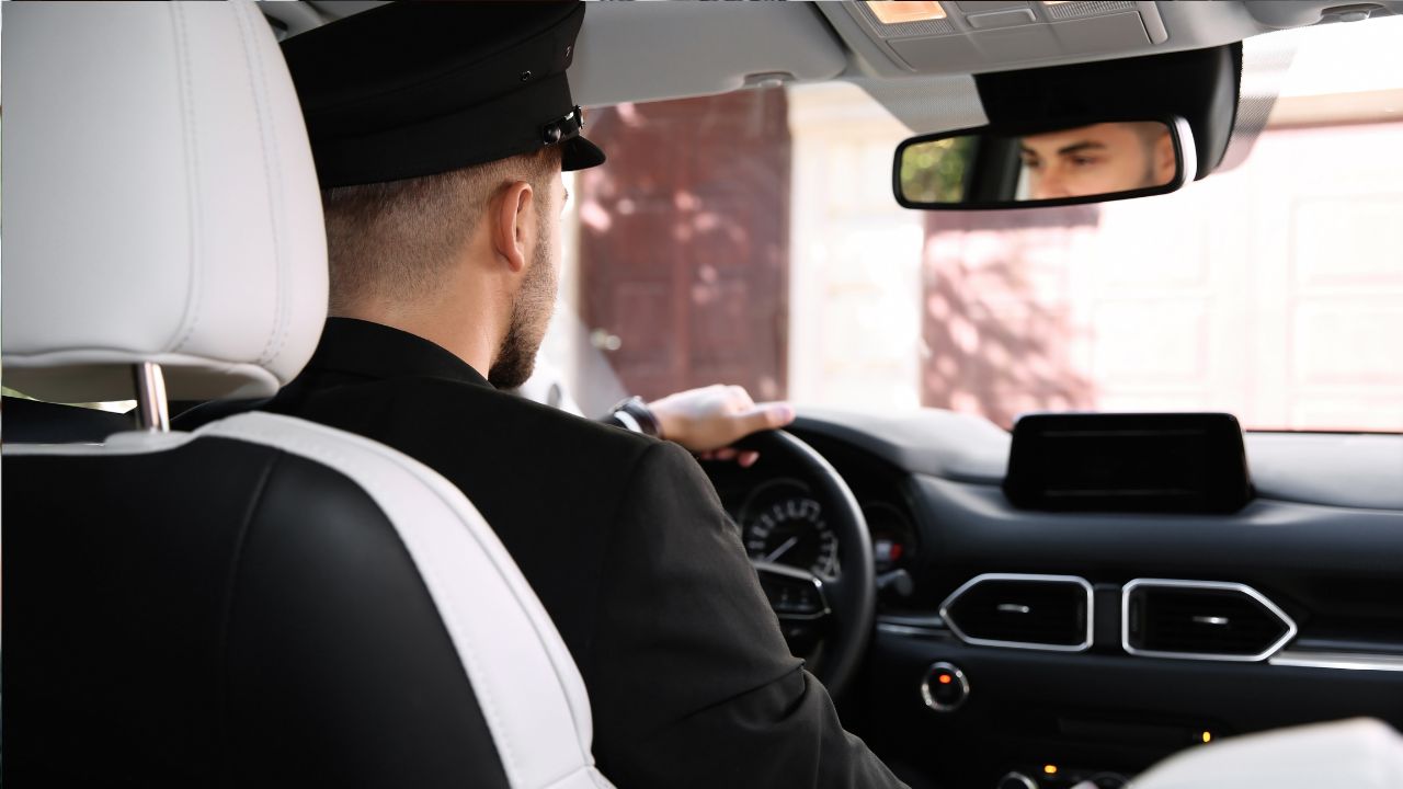 Šofér v uniforme riadi auto, pohľad zo zadného sedadla na palubnú dosku a spätné zrkadlo.
