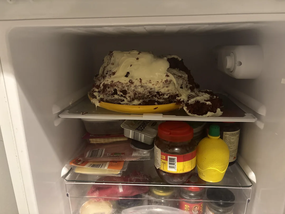 Pokazená torta v chladničke na žltom tanieri, obklopená rôznymi potravinami a ingredienciami na policiach.