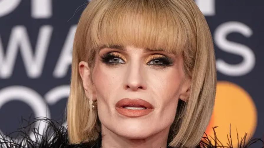 Osoba s krátkym blond účesom a výrazným líčením na červenom koberci počas slávnostného podujatia.