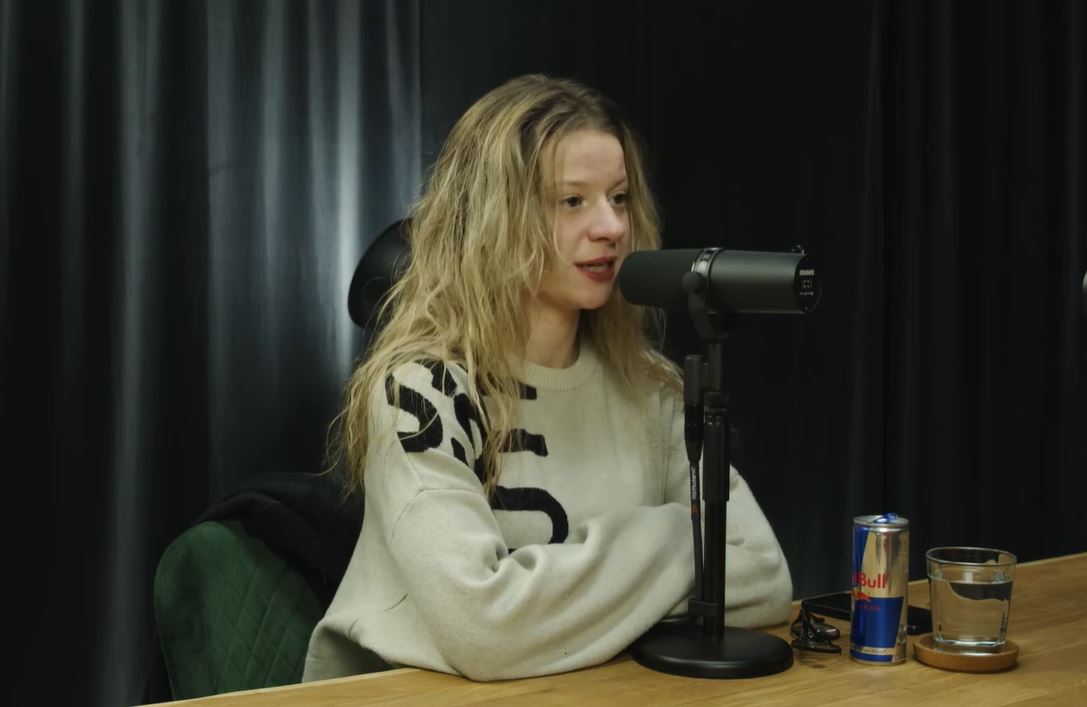 Osoba pri mikrofóne v nahrávacom štúdiu s nápojom na stole počas podcastu.