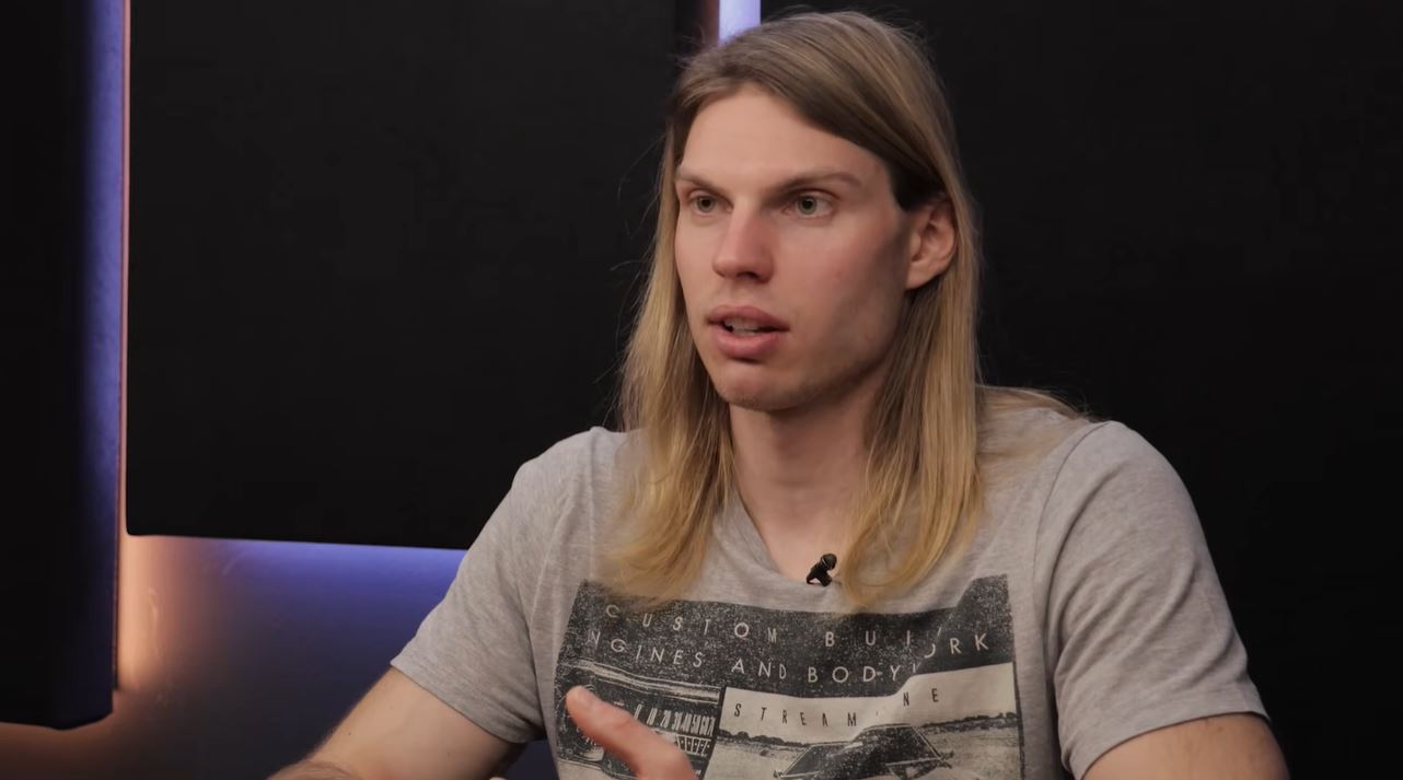 Osoba s dlhými blond vlasmi hovorí pred čiernymi zvukovými panelmi počas diskusie.