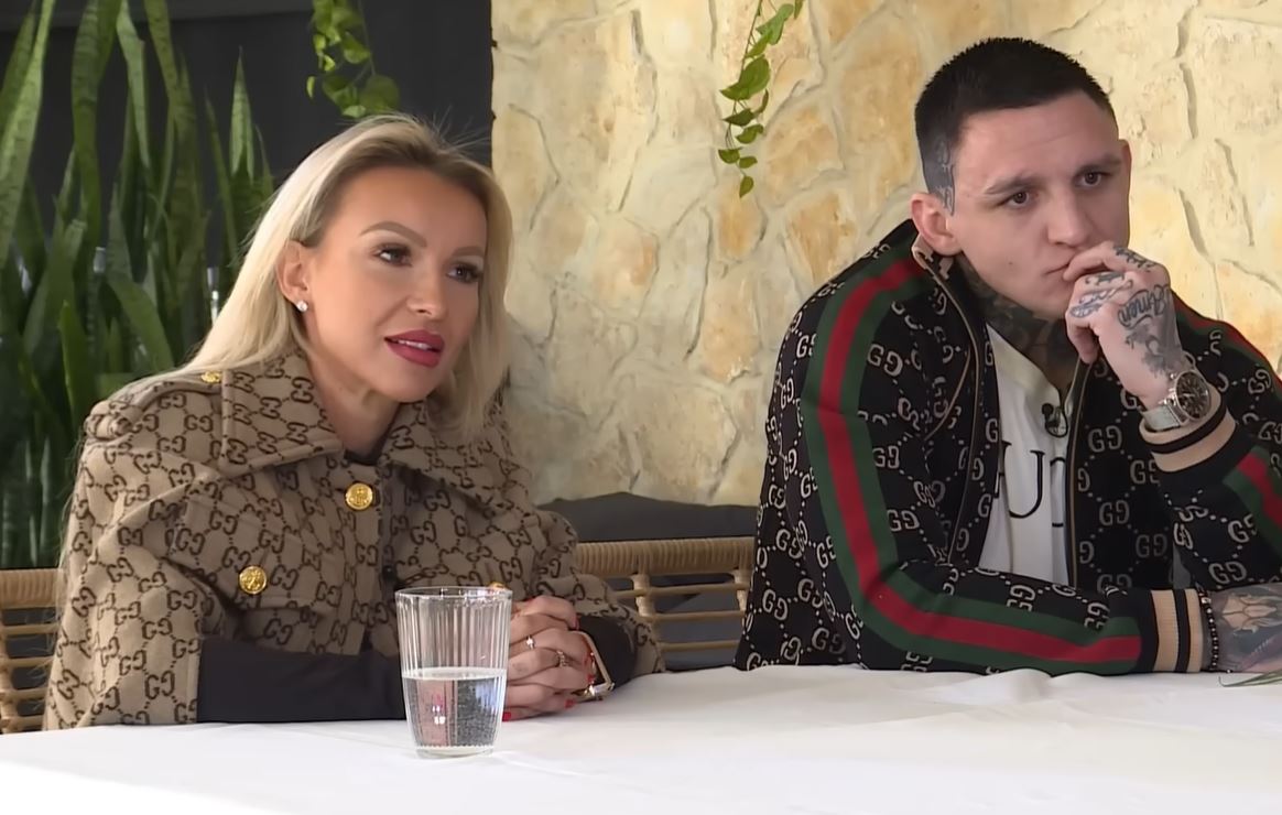 Dvaja ľudia sedia pri stole, diskutujú, oblečení v značkovom oblečení s logom Gucci na svetlom pozadí.
