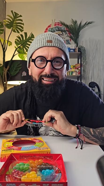 Muž s okuliarmi a čiapkou sedí pri stole, drží žuvacie cukríky a smeje sa. Na stole sú rôzne farebné sladkosti.