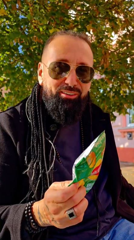 Muž v slnečných okuliaroch, sediaci pod stromom, drží balík chipsov a usmieva sa na slnečnom dni.