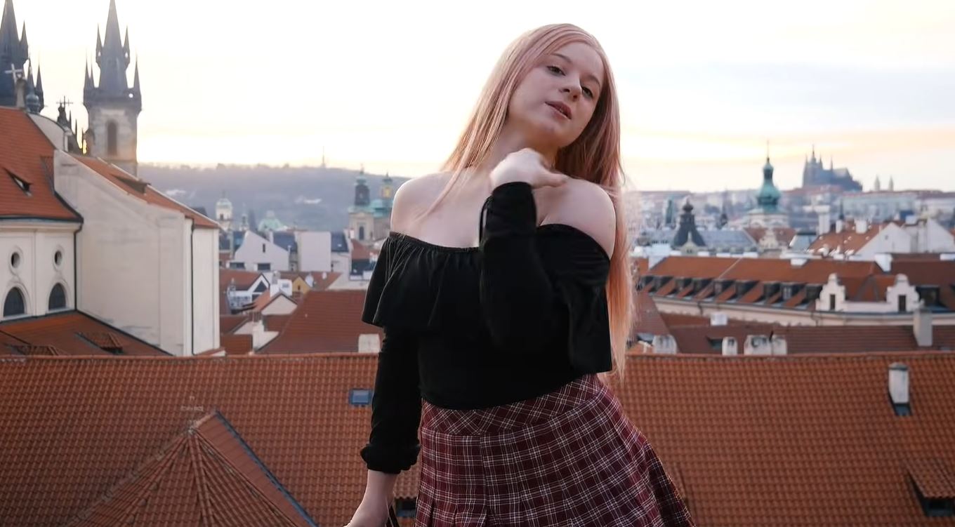 Osoba na streche s výhľadom na Prahu; večerný západ slnka, červené strechy a historická architektúra na pozadí.