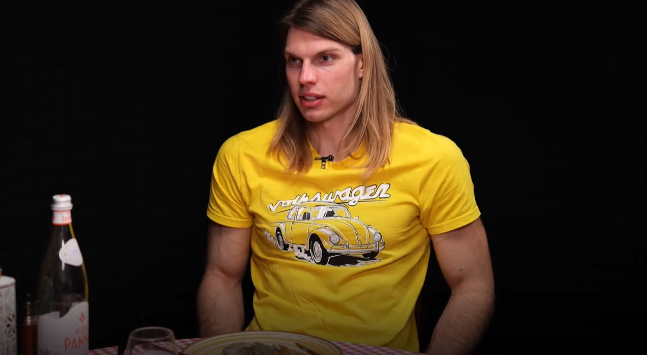 Muž v žltom tričku s motívom vozidla Volkswagen sedí pri stole s fľašou vody a jedlom.