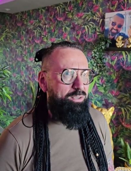 Obývačka so zelenou tapetou a muž s dlhými vlasmi a bradou nosí okuliare, v pozadí rámik s obrázkom.