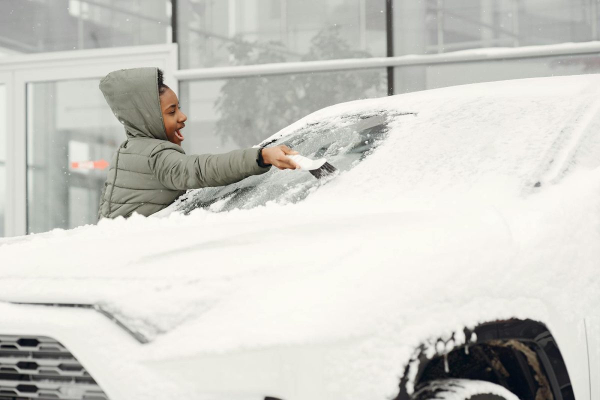 A woman in a gray coat clears snow from a car windshield on a cold winter day.