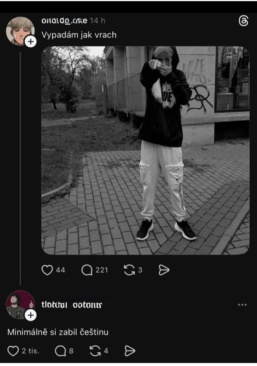Mladý človek v mikine a teplákoch pózuje vonku pri budove, s graffiti na pozadí.