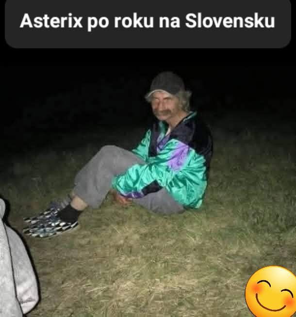 Muž v barevné bundě sedí na trávě v noci s vtipným žltým smajlíkom v rohu.