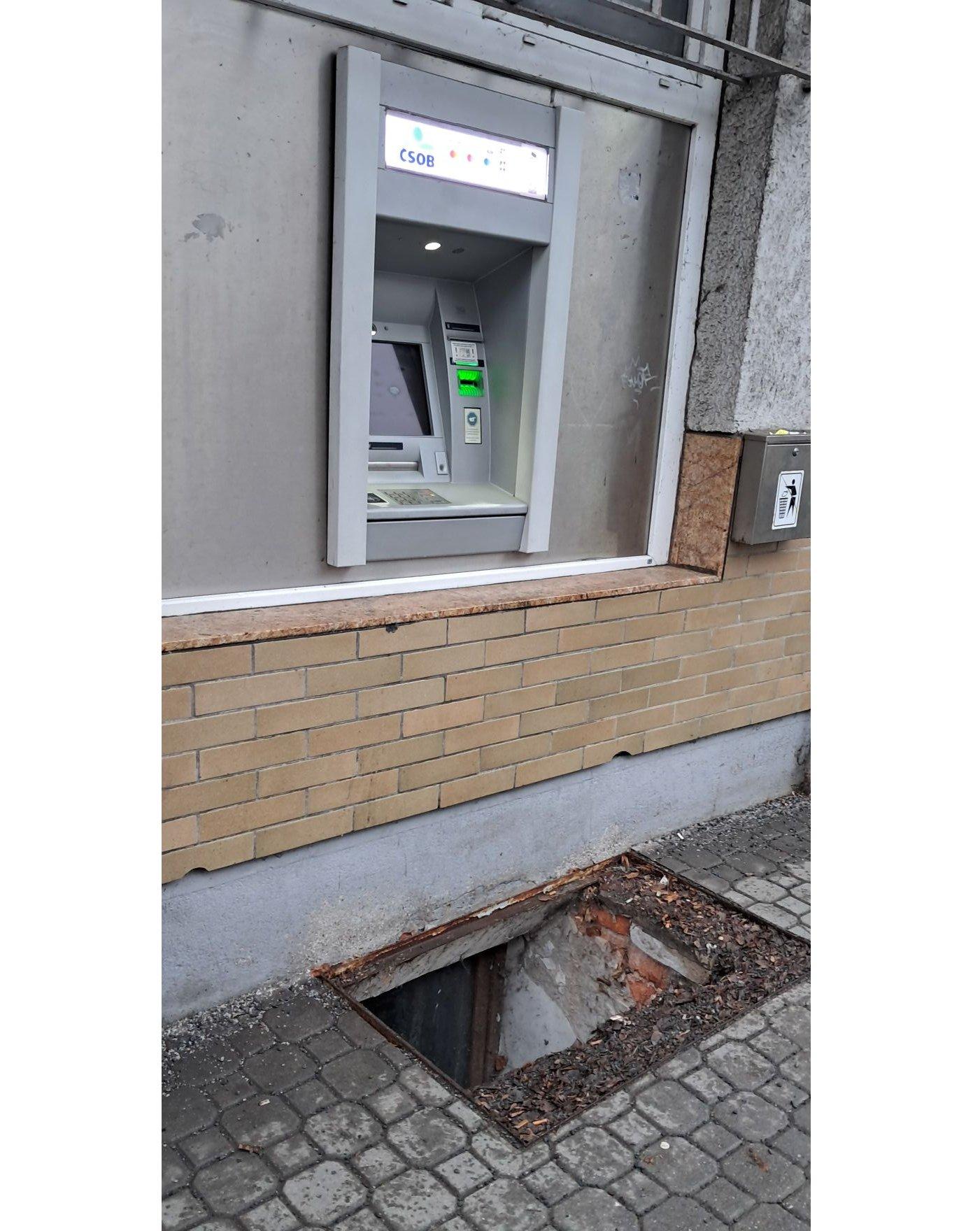 Bankomat ČSOB, nad poškodeným chodníkom s otvorom, v centre mesta.