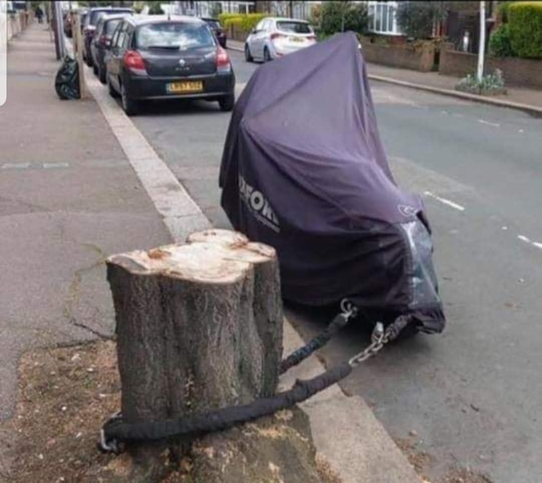 Motorka zamaskovaná plachtou pripevnená reťazou k pňu na ulici plnej áut.