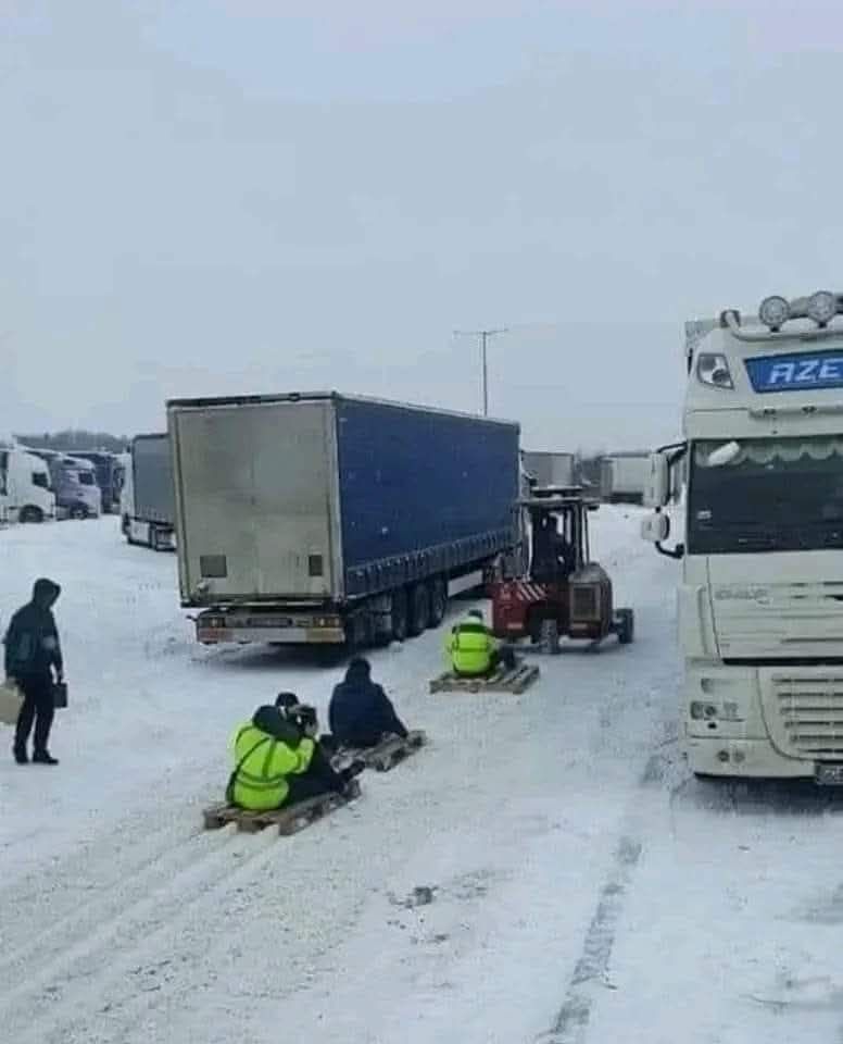 Ľudia na saniach ťahaní vysokozdvižným vozíkom po zasneženej ceste plnej kamiónov.