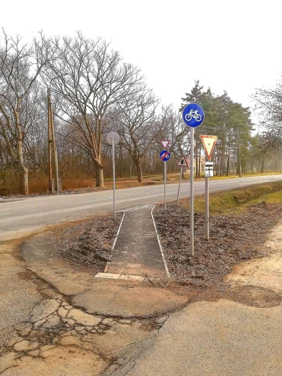 Cyklistický chodník na križovatke so značkami, okolie s holými stromami a zamračenou oblohou.