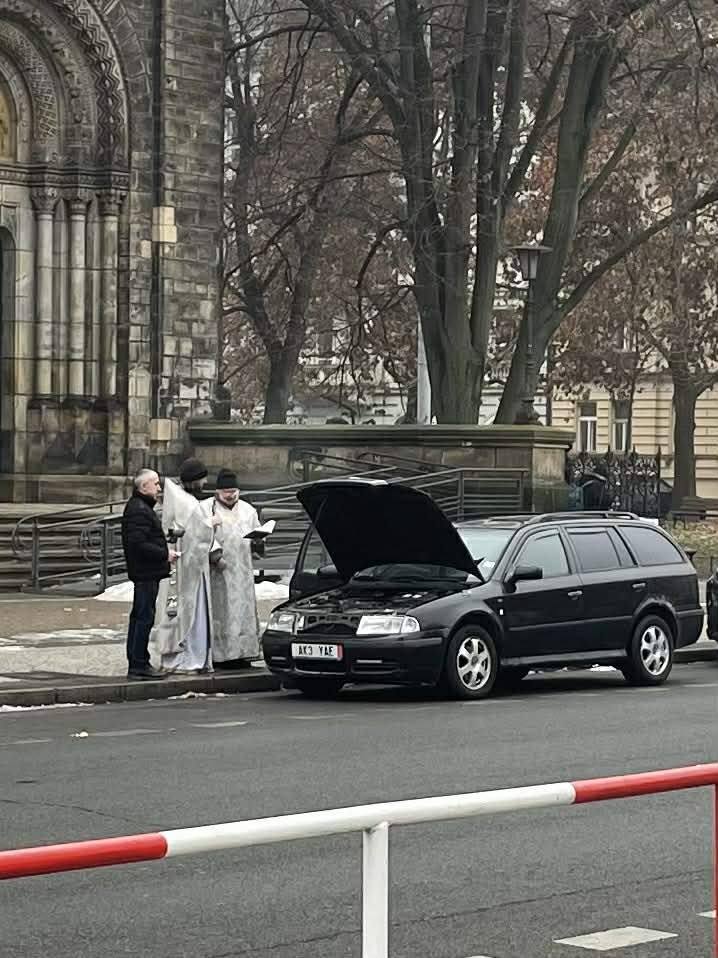 Skupina ľudí pri zaparkovanom aute s otvorenou kapotou pred historickou budovou v zime.