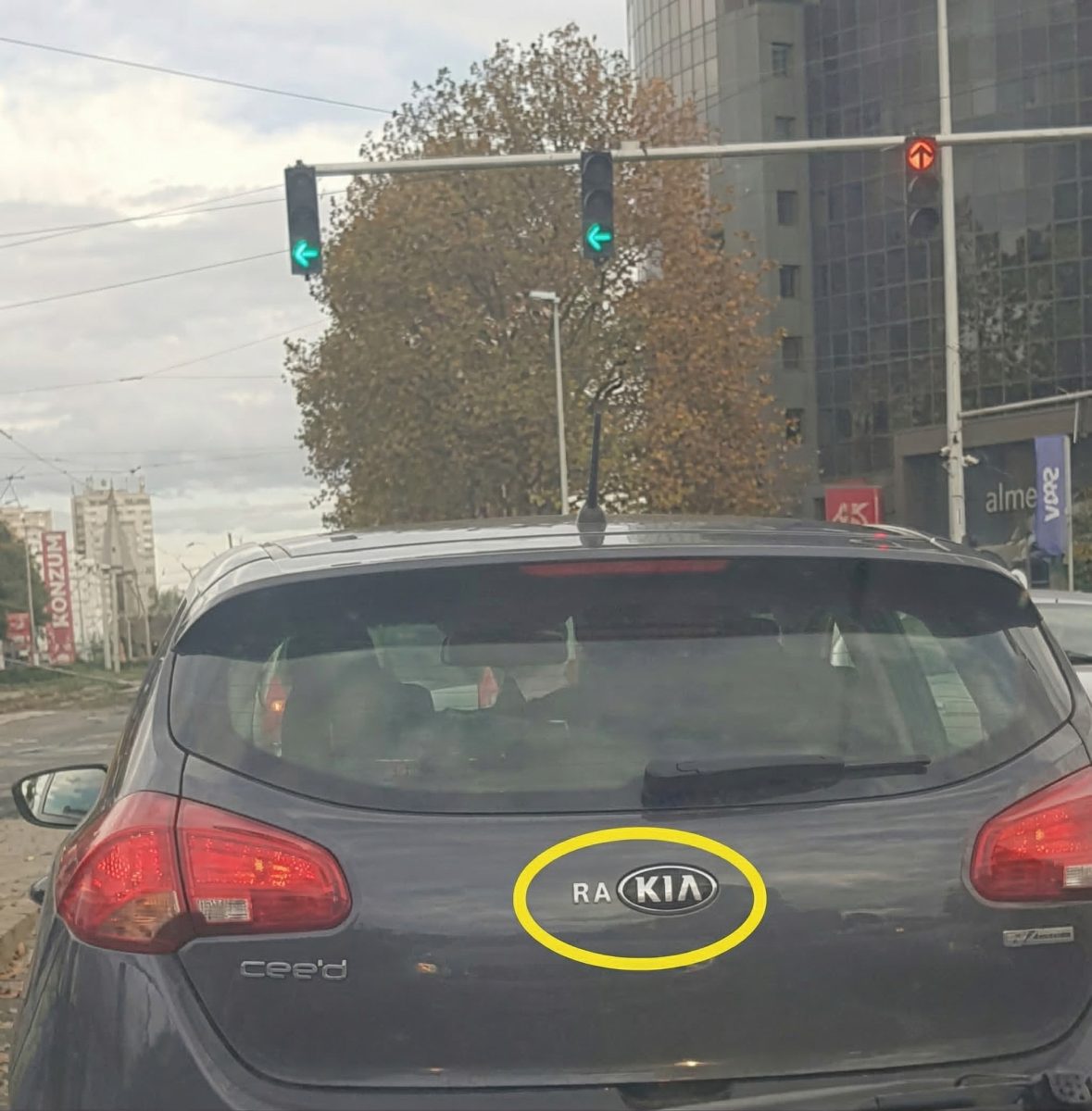 Auto značky Kia cee'd na križovatke so semaforom, pod jesenným nebom, s budovou v pozadí.