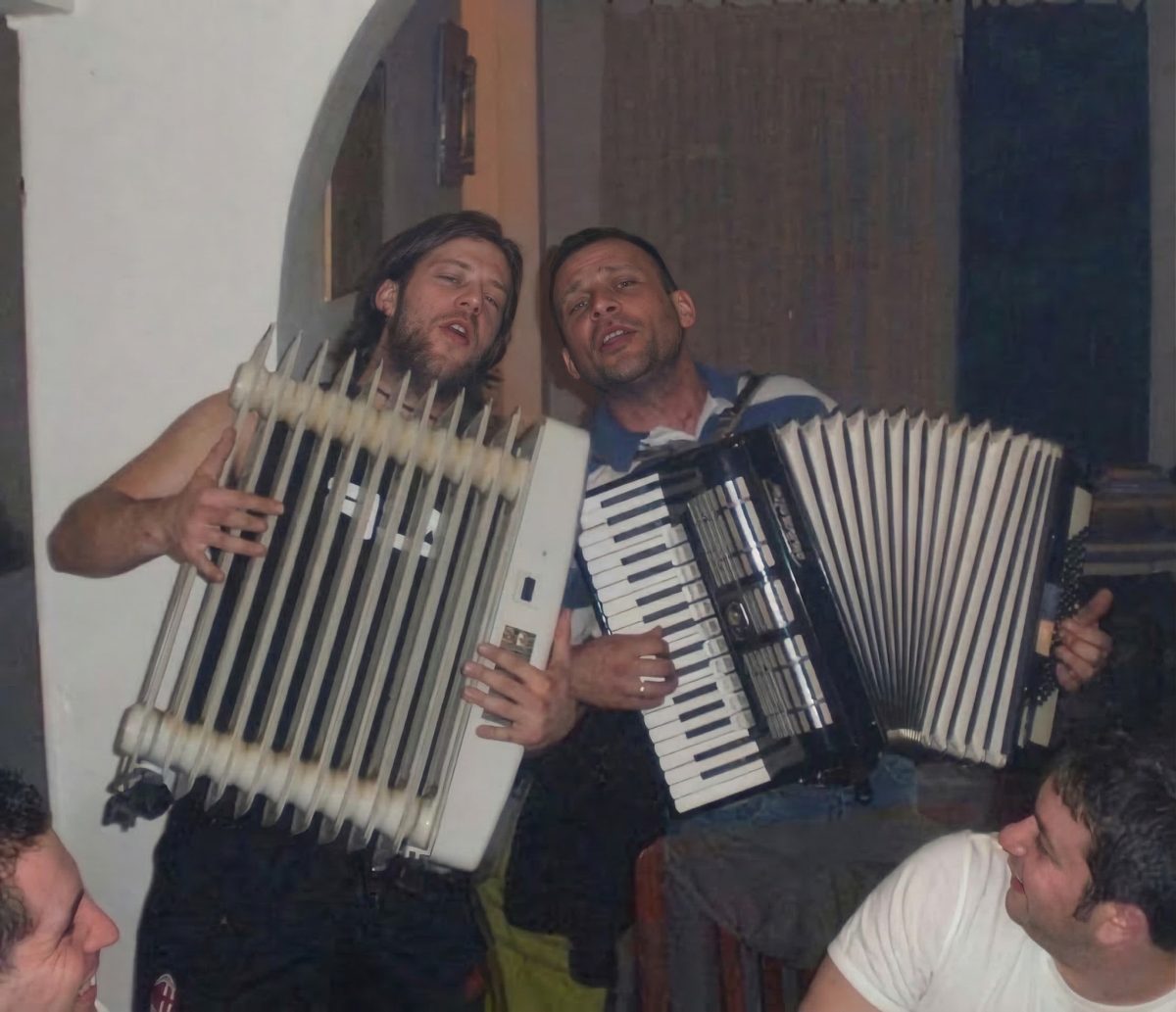 Dvaja muži v interiéri hrajú na harmonikách, jeden improvizuje s radiátorom, okolo nich sedí skupina ľudí.